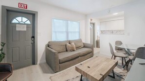 A modern living room with a grey door marked 'EXIT', a beige sofa, a wooden coffee table, and a dining area with a white table and grey chairs.