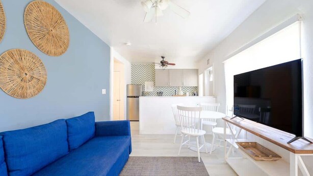 A modern living room with a blue sofa, a dining area with white chairs, a kitchen with white cabinets, and a large TV on a wooden stand.