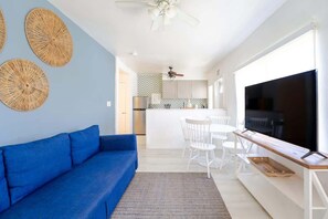 Een moderne woonkamer met een blauwe bank, een eethoek met witte stoelen, een keuken met witte kastjes en een grote televisie op een houten dressoir.