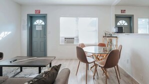 A modern living room with a glass dining table, two sofas, and a coffee table. There are two exits marked with red signs, and a window with an air conditioning unit.