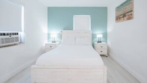 A bedroom with a white bed, two bedside tables with lamps, a window with blinds, and a wall-mounted air conditioner. - Sunset Cove In Hollywood FL, FREE Parking (Hollywood)