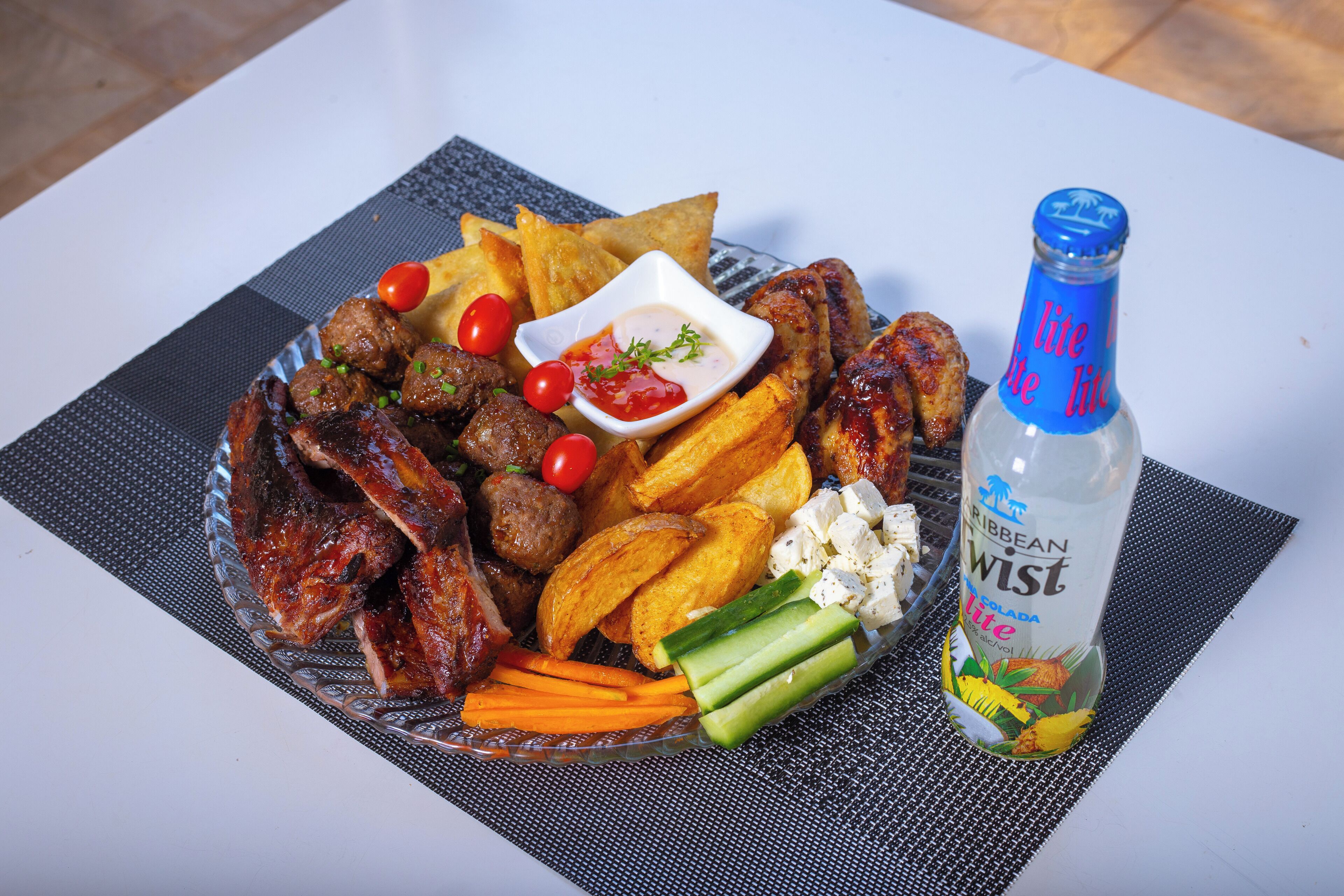 Un plato de vidrio con diversos alimentos, incluyendo albóndigas, papas fritas y verduras, además de una botella de Caribbean Twist sobre una mesa.