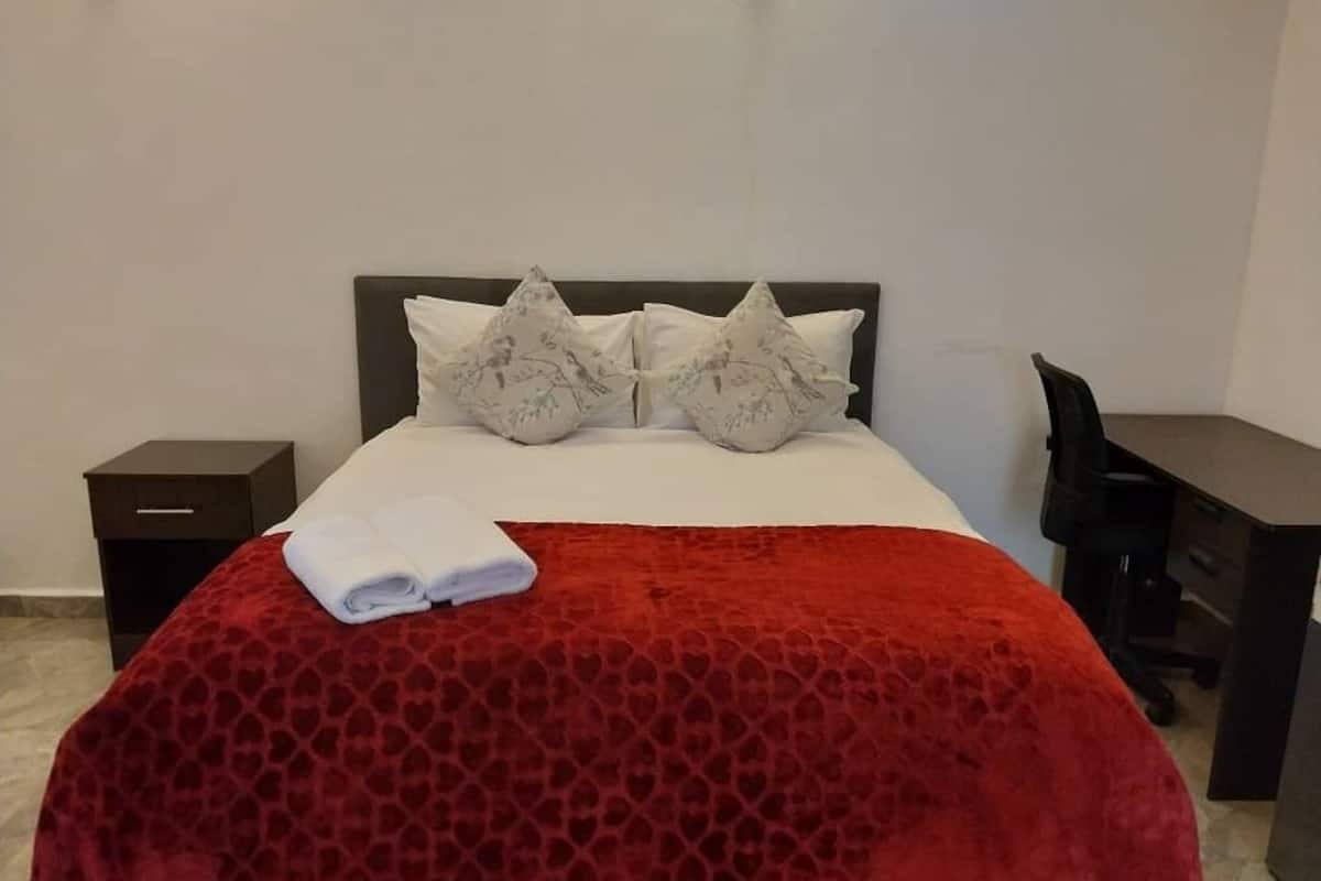 A neatly made bed with a red patterned bedspread and two pillows, a small bedside table, and a desk with a chair.