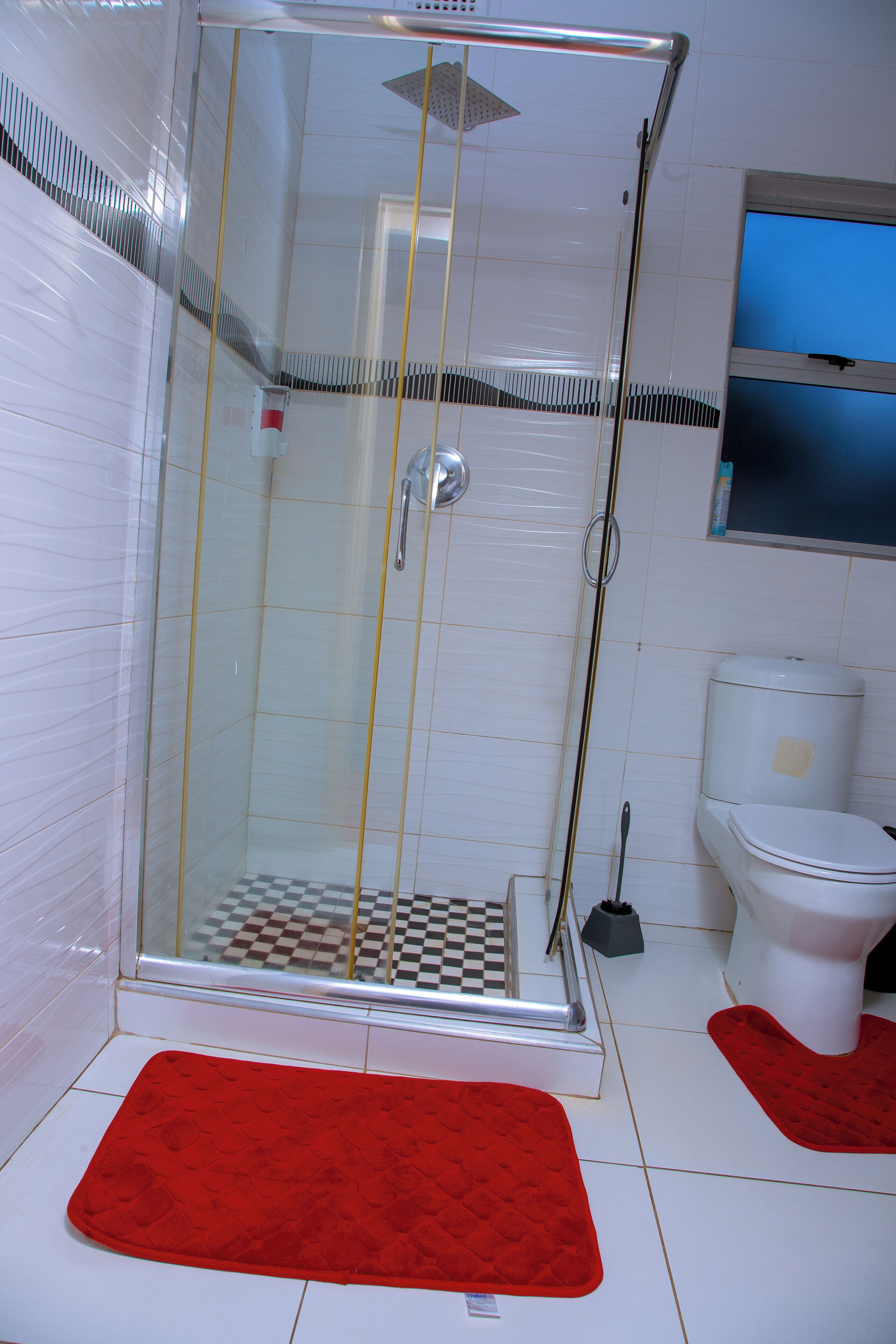 A glass-enclosed shower with a checkered floor mat, a red bath mat, and a toilet with a white tank.