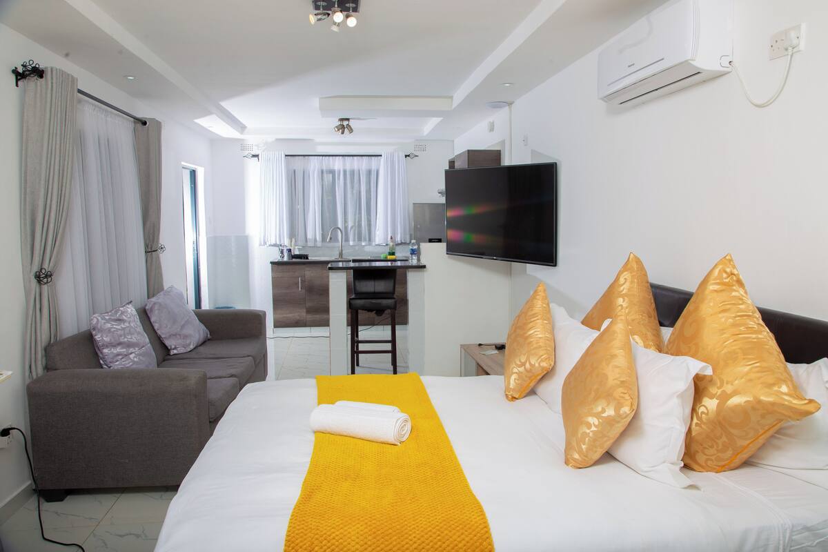 A hotel room with a bed, a sofa, a TV, and a kitchenette in the background.