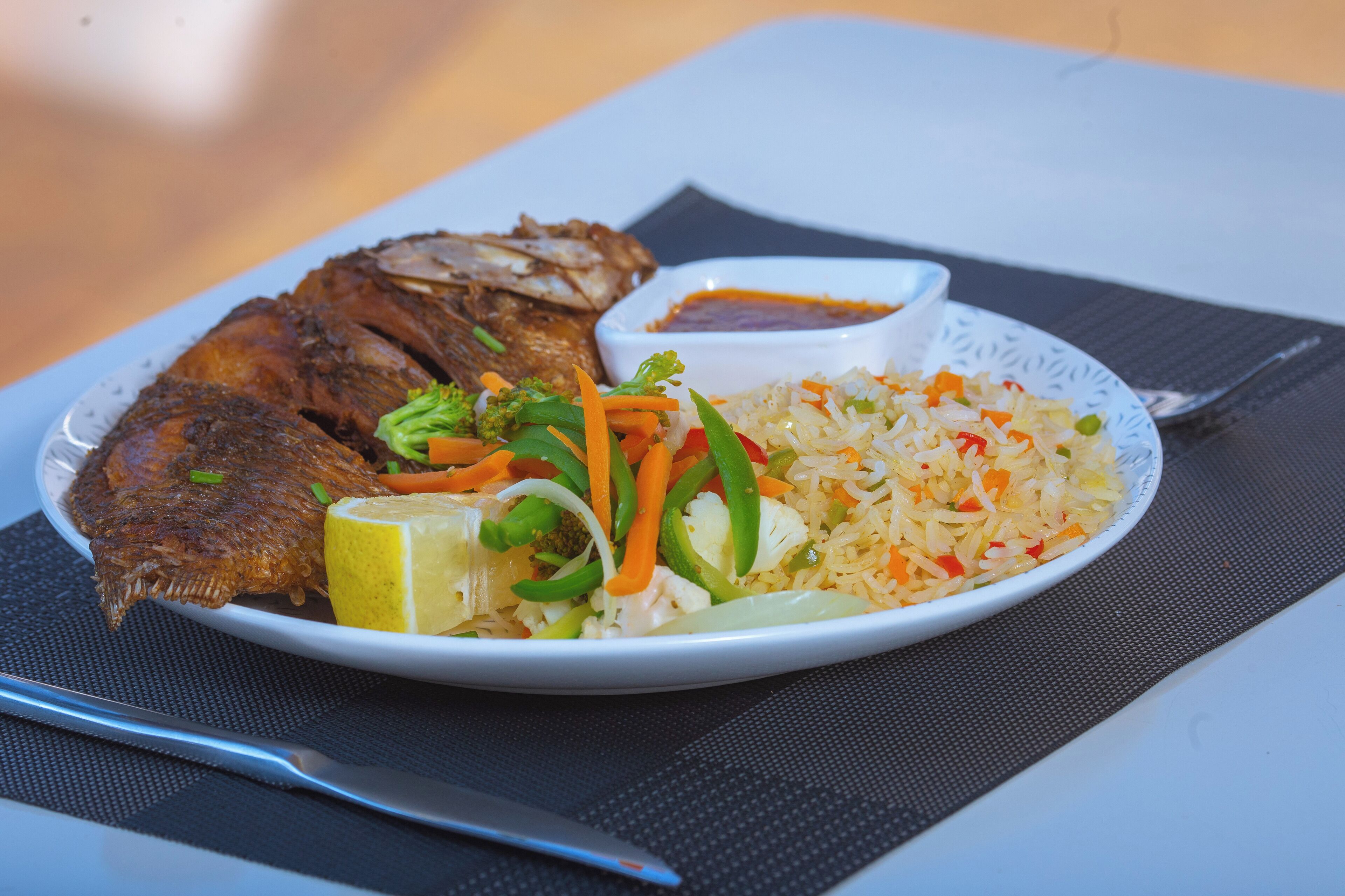 A plate of fried fish, rice, and vegetables with a lemon wedge and a small bowl of sauce.