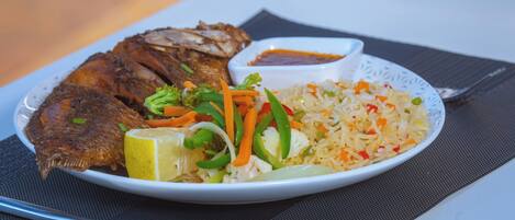 A plate of fried fish, rice, and vegetables with a lemon wedge and a small bowl of sauce.