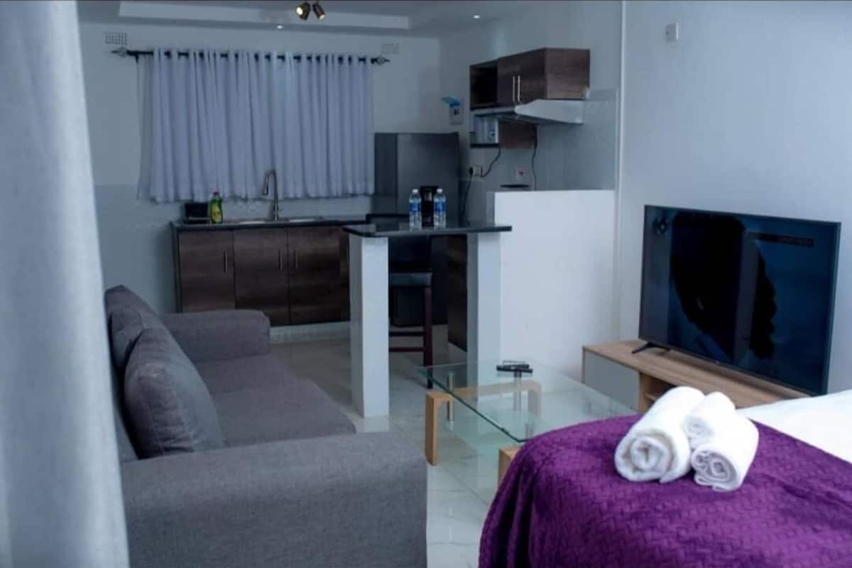 A modern hotel room with a grey sofa, a small kitchen area with a microwave, a glass table, and a TV on a wooden stand.