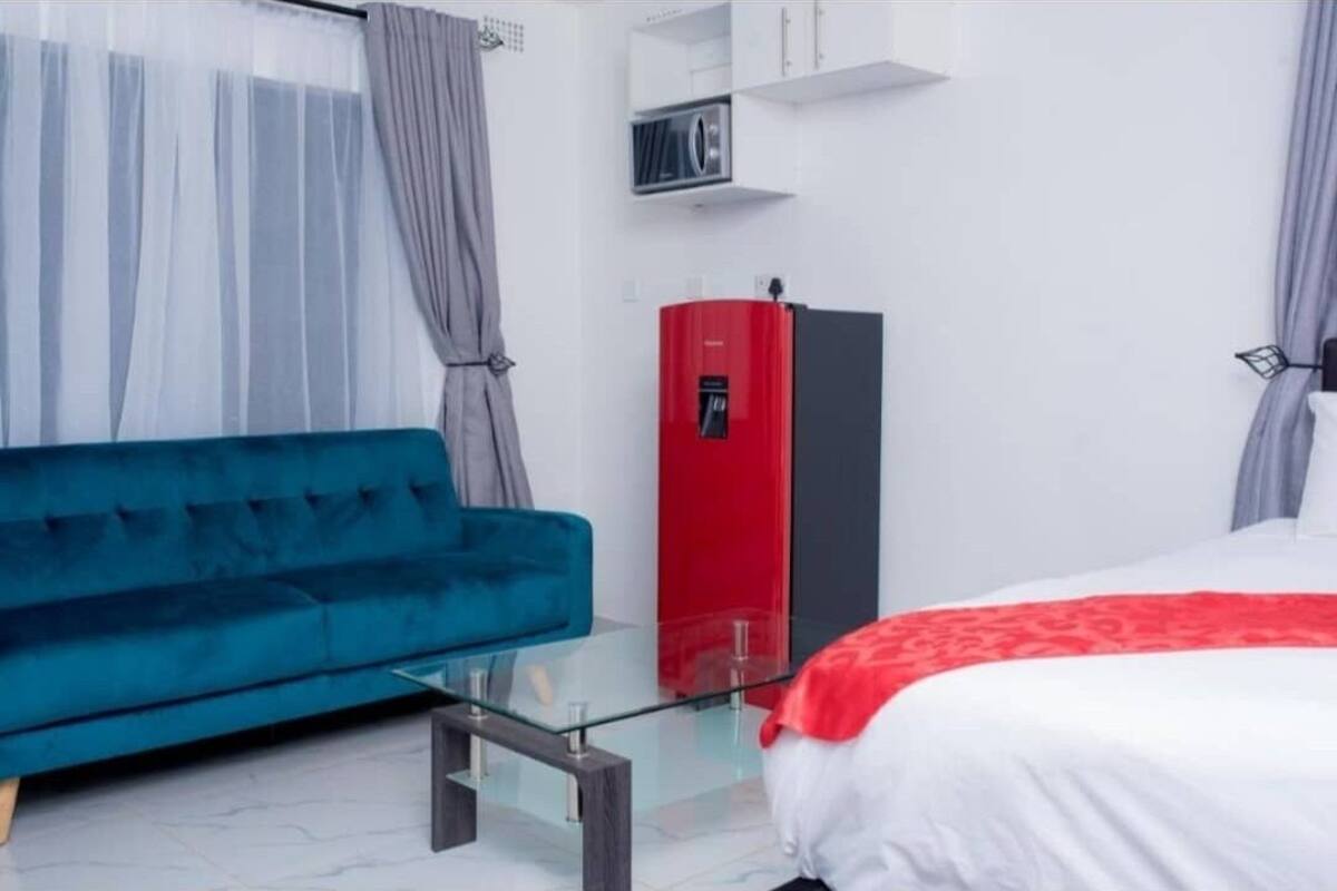 A modern hotel room with a teal sofa, a glass coffee table, a bed with a red and white bedspread, and a red minibar.