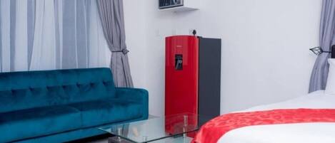 A modern hotel room with a teal sofa, a glass coffee table, a bed with a red and white bedspread, and a red minibar.