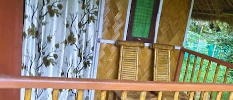 A wooden balcony with a thatched roof, a wooden railing, and a view of a bamboo structure.