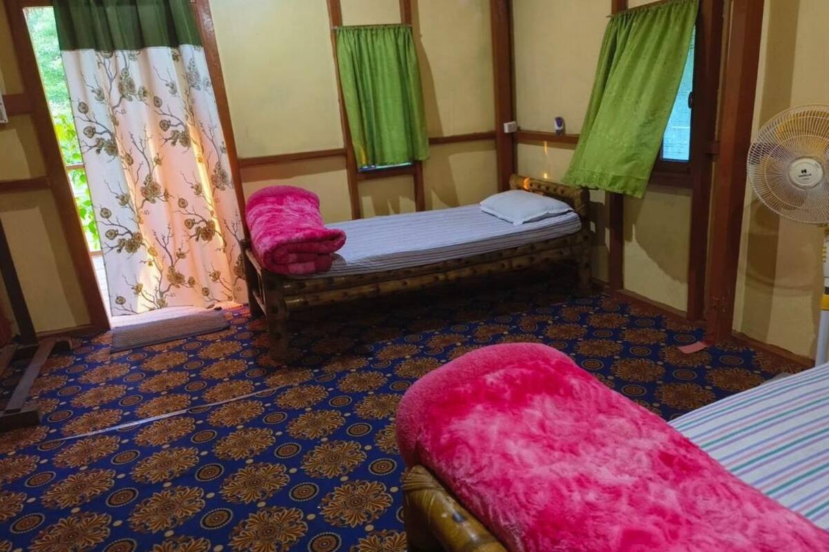 A room with a bed, a chair, a standing fan, and a window with curtains.