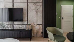 A modern living room with a flat-screen TV mounted on a wall with a floral pattern, a black entertainment unit, a round table, and a green chair. - Symphony Atal City Square (Wroclaw)