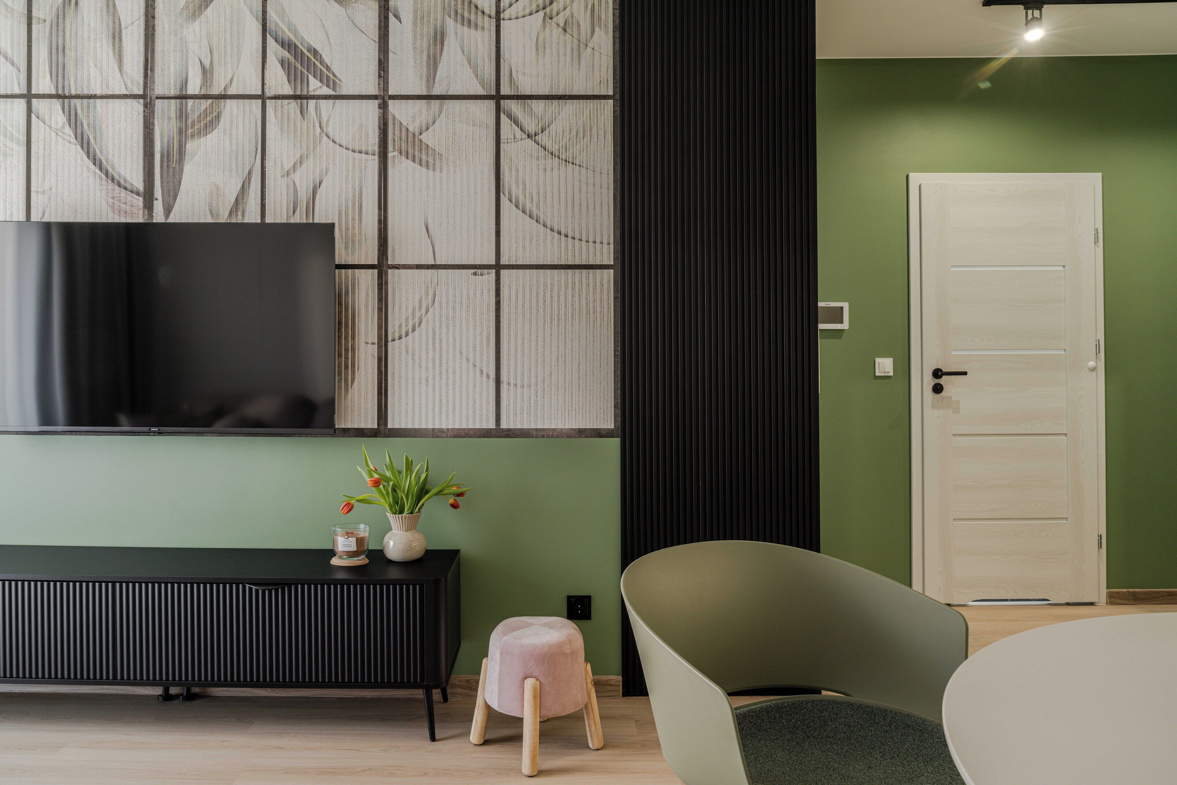 Una sala de estar moderna con un televisor de pantalla plana, una mesa consola negra, un taburete rosa, un sillón verde y una puerta blanca.