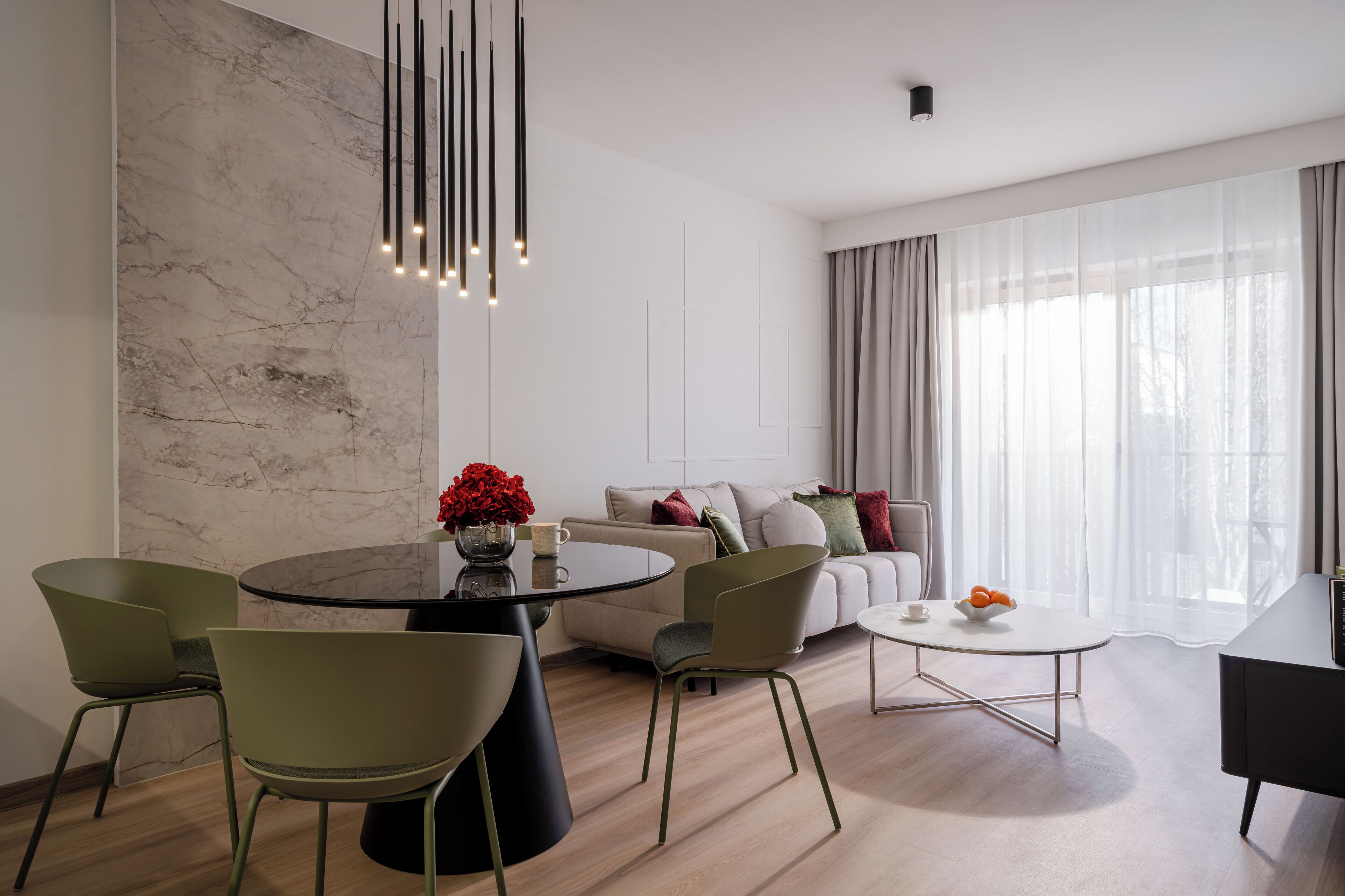 Una sala de estar moderna con un muro decorativo de mármol, una mesa redonda de comedor con sillas, un sofá con cojines y un ventanal con cortinas translúcidas.