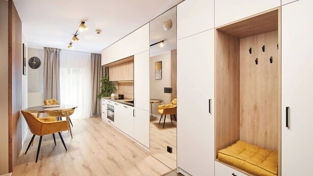A modern kitchen with white cabinets and a wooden countertop. There is a dining area with a yellow chair and a table. - Symphony Atal City Square (Wroclaw)