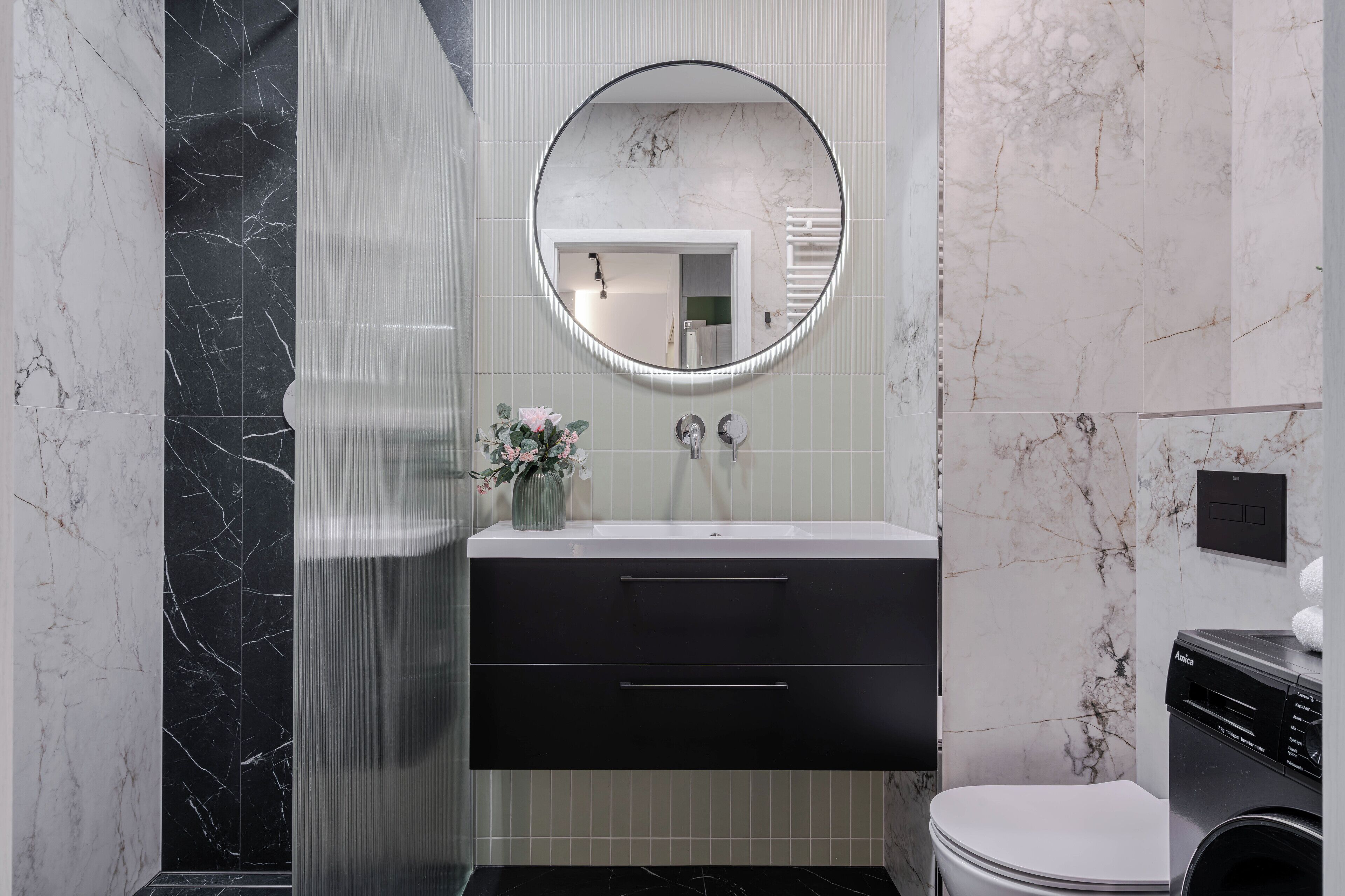 A modern bathroom with a black marble vanity, a round mirror, a toilet, and a washing machine.