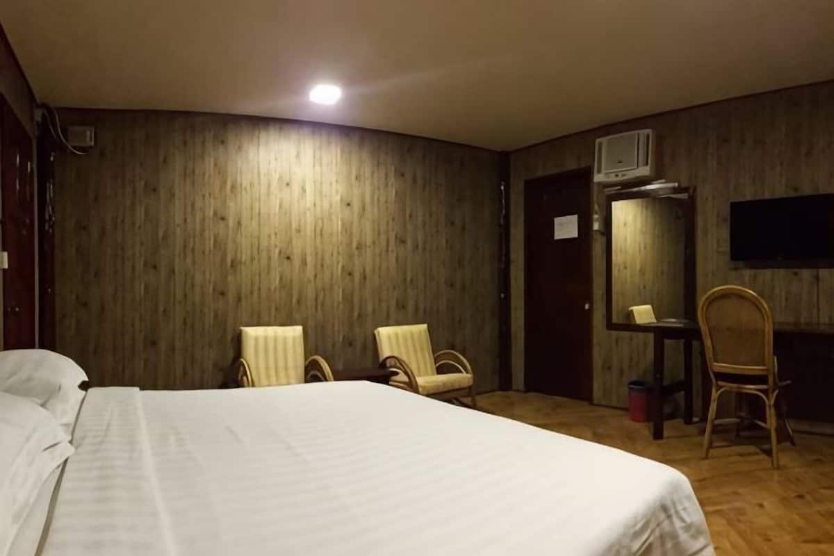 A hotel room with a bed, two chairs, a desk, a mirror, and an air conditioning unit.