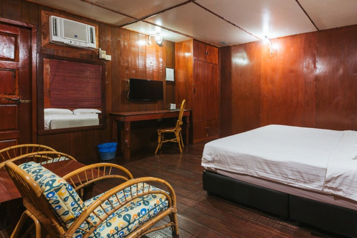 A room with a bed, a desk, a television, and wicker chairs.