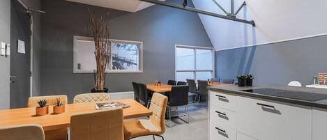 Une cuisine moderne dotée d’un grand îlot central, d’un coin repas avec une table en bois et des chaises rembourrées, et d’une vue sur un chantier de construction par la fenêtre.
