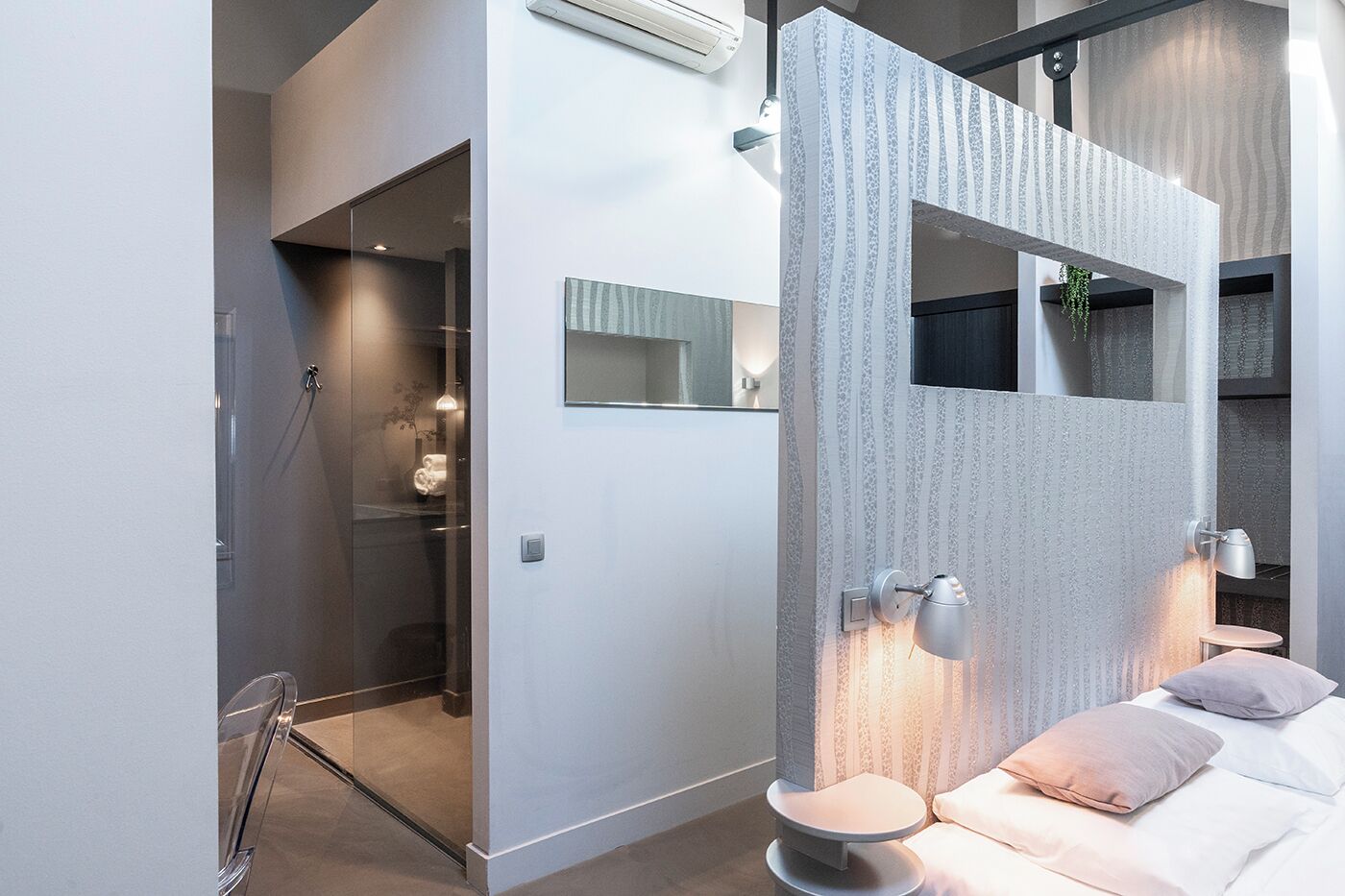 A modern bathroom with a glass shower enclosure, a vanity with a mirror, and a bedside table with a lamp.