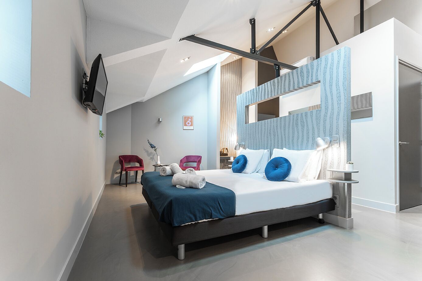 A modern bedroom with a large bed, two red chairs, a TV mounted on the wall, and a mirror with a patterned frame.