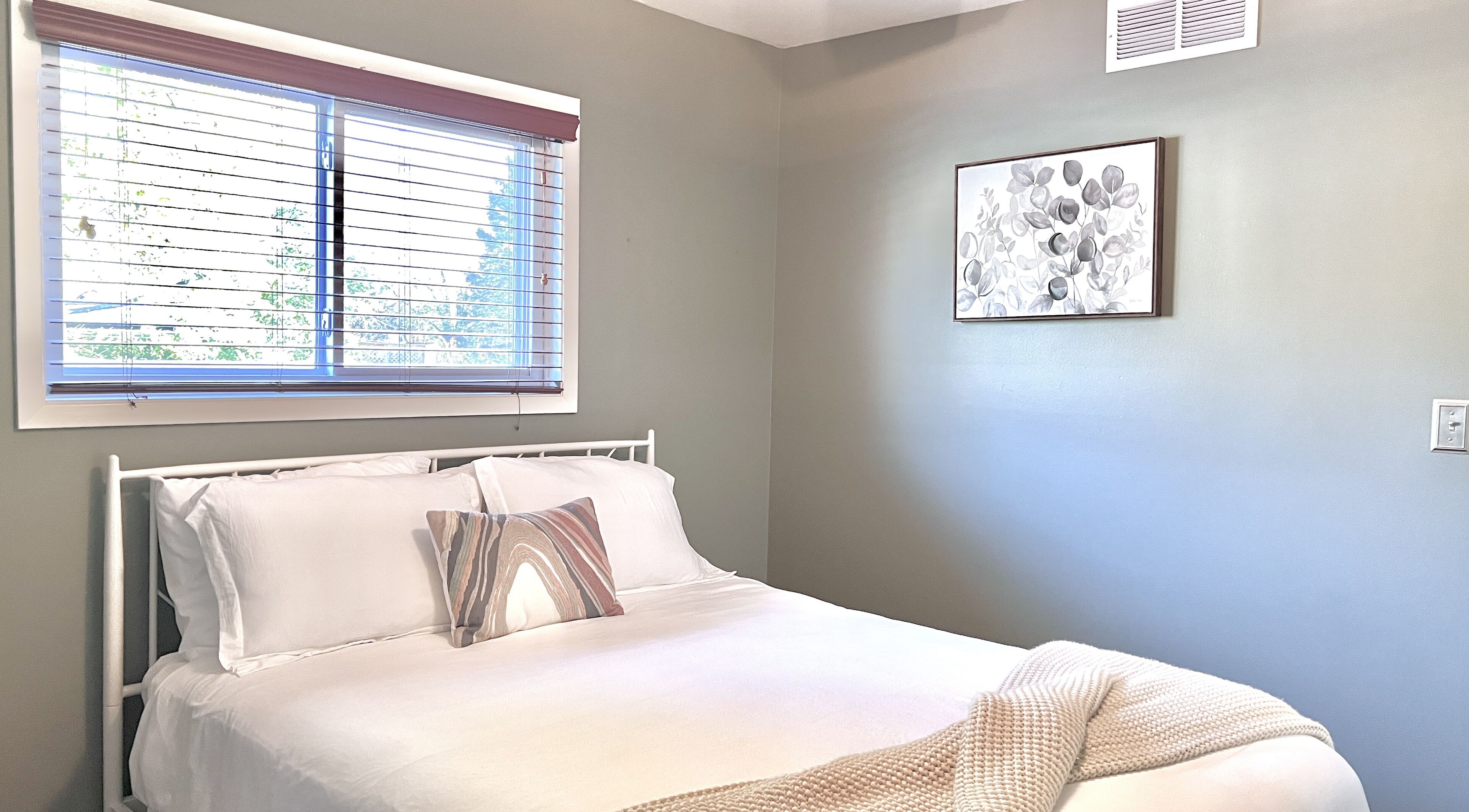 Un lit bien fait, avec du linge de lit blanc et un seul coussin à motifs, une fenêtre avec des stores et une œuvre d’art florale encadrée accrochée au mur.