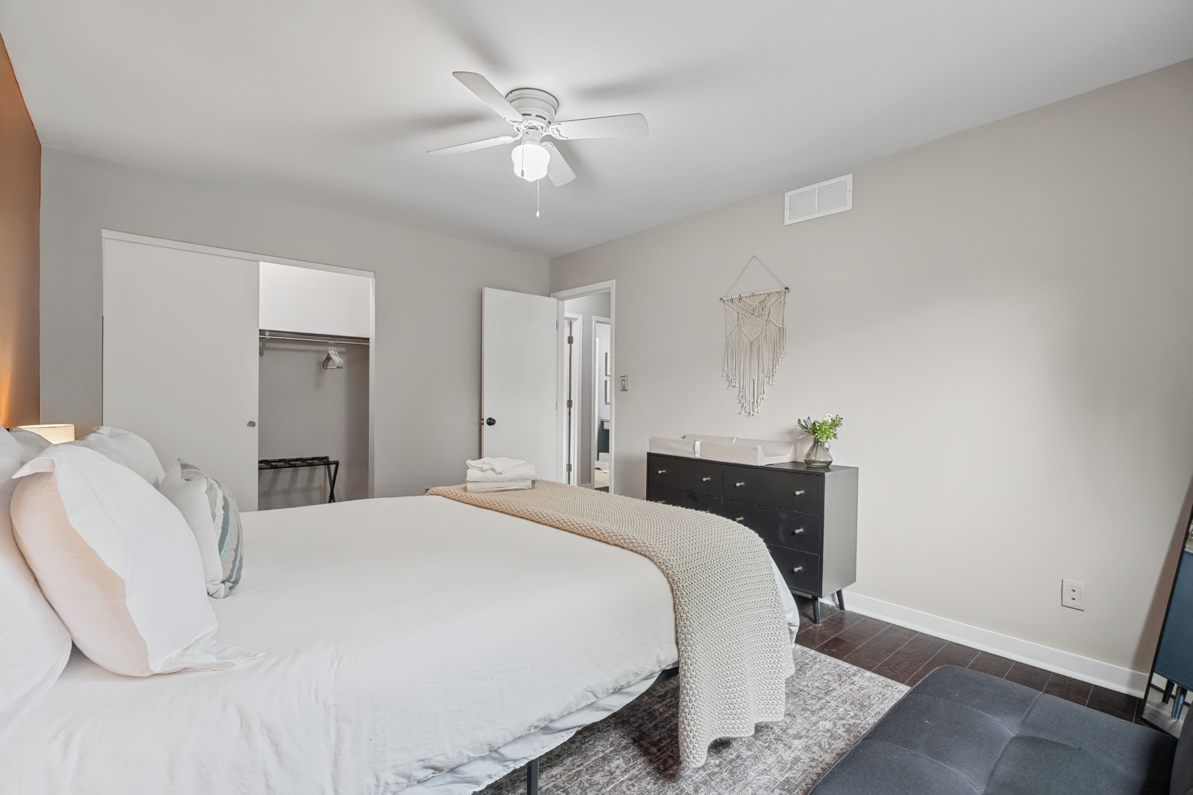 A bedroom with a bed, a dresser, a ceiling fan, and a wall-mounted macrame decoration.