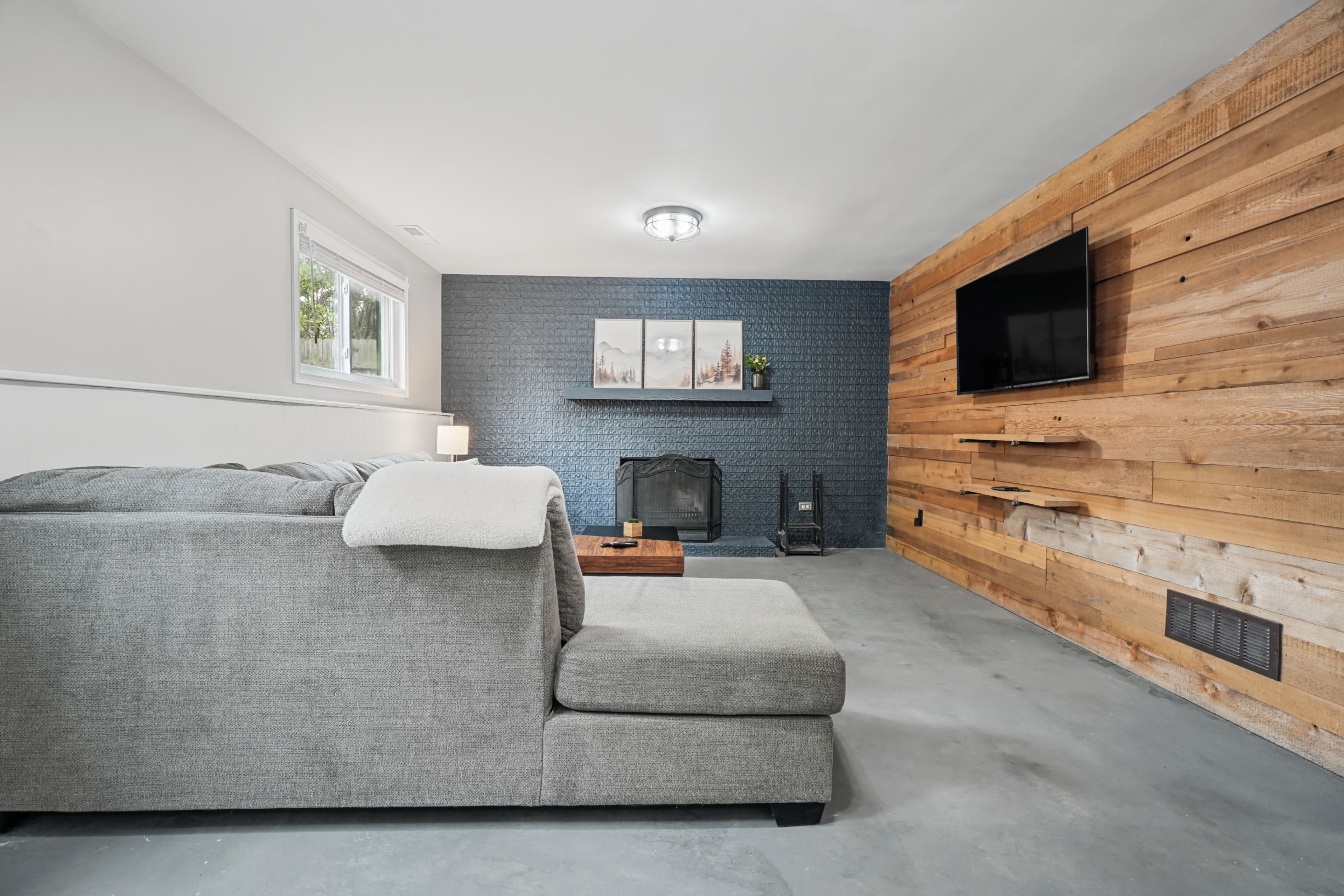 A modern living room with a grey sectional sofa, a fireplace, and a flat-screen TV mounted on a wooden wall.