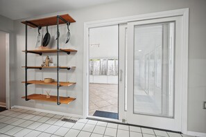 A kitchen with a white tiled floor, a black metal shelving unit with kitchen utensils, and a sliding glass door leading to an outdoor area.