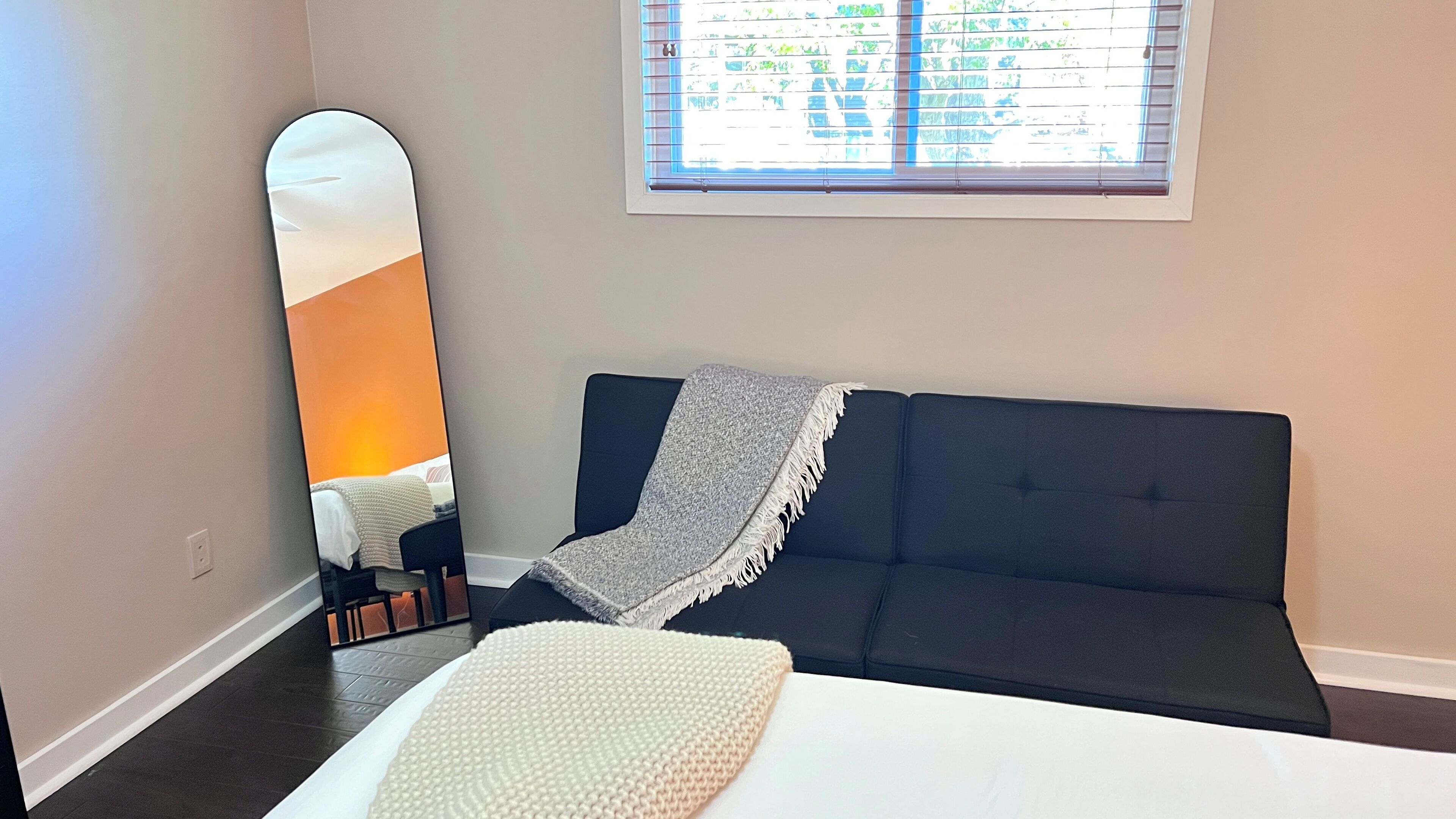 Une chambre à coucher avec un canapé de couleur sombre, un lit blanc et un grand miroir qui reflète une fenêtre munie de stores.