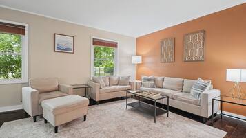 A living room with two sofas, a coffee table, and a rug. There are two windows with blinds, a painting on the wall, and a floor lamp.