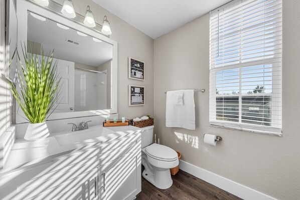 A modern bathroom with a white vanity, a large mirror, a toilet, and a window with blinds.