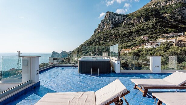 A rooftop pool with lounge chairs, a hot tub, and a view of the sea and mountains.