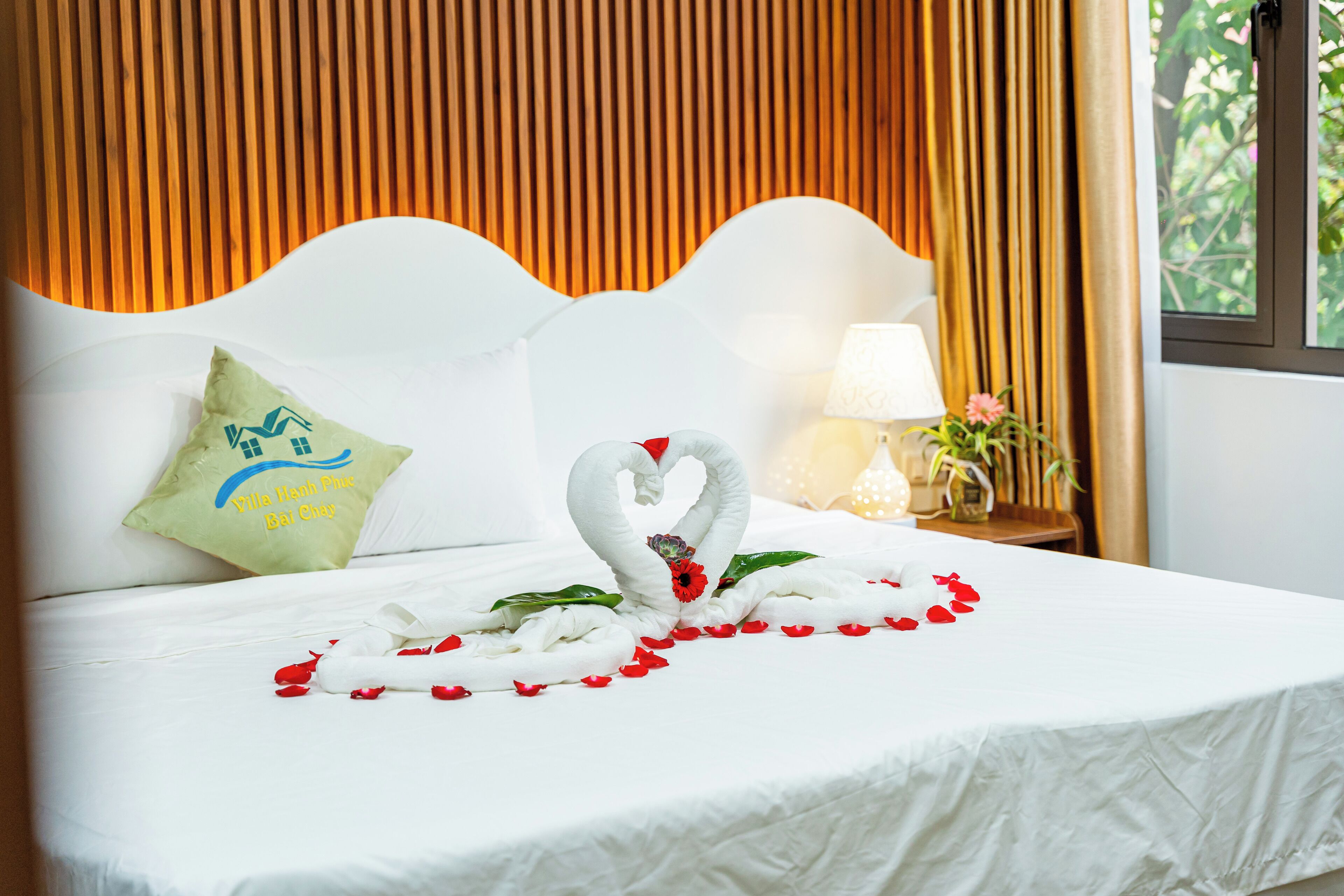 A neatly made bed with a swan-shaped towel and rose petals, a pillow with a hotel logo, and a lamp on a bedside table.