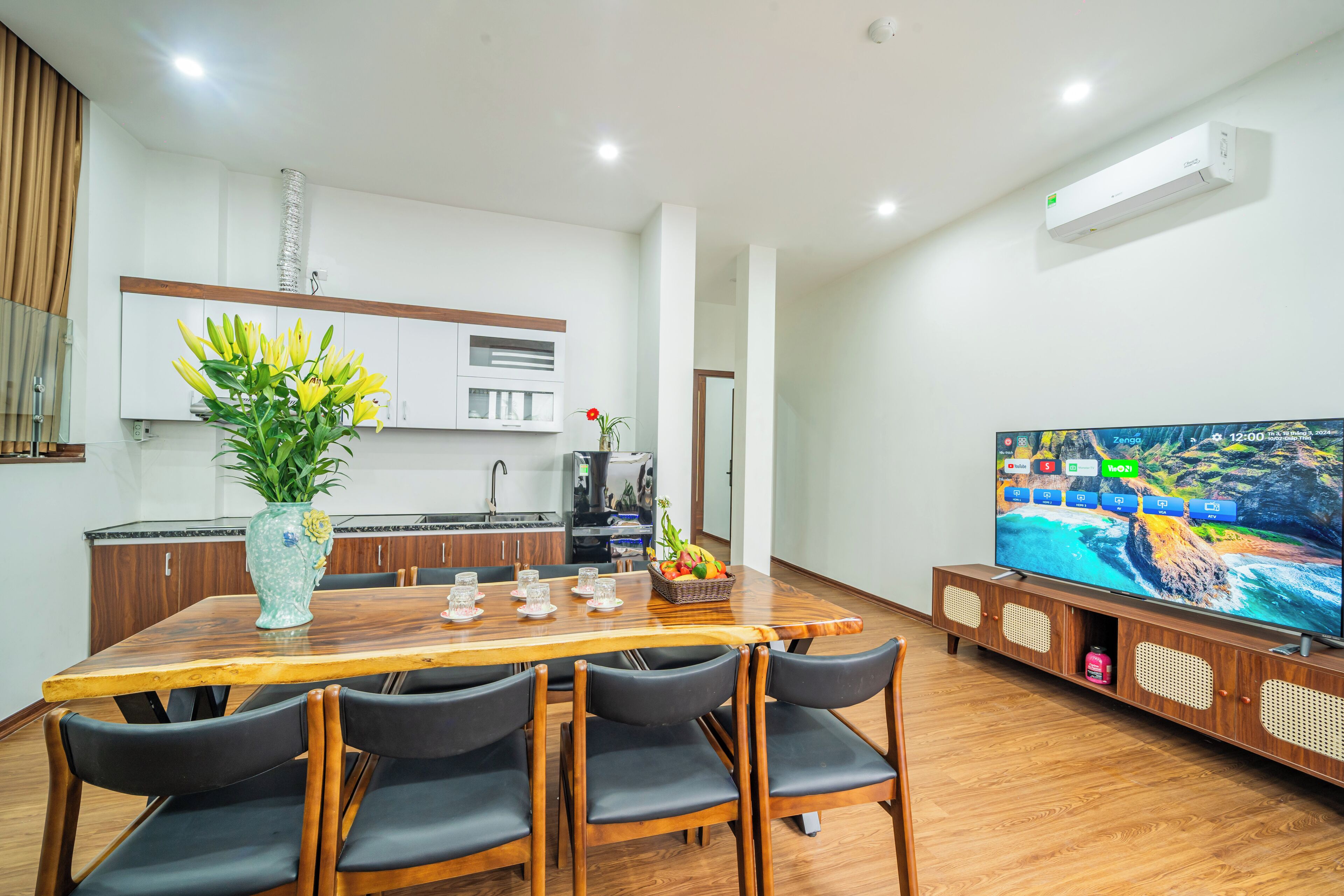 A modern living room with a wooden dining table, black chairs, a TV, and a kitchen area in the background.
