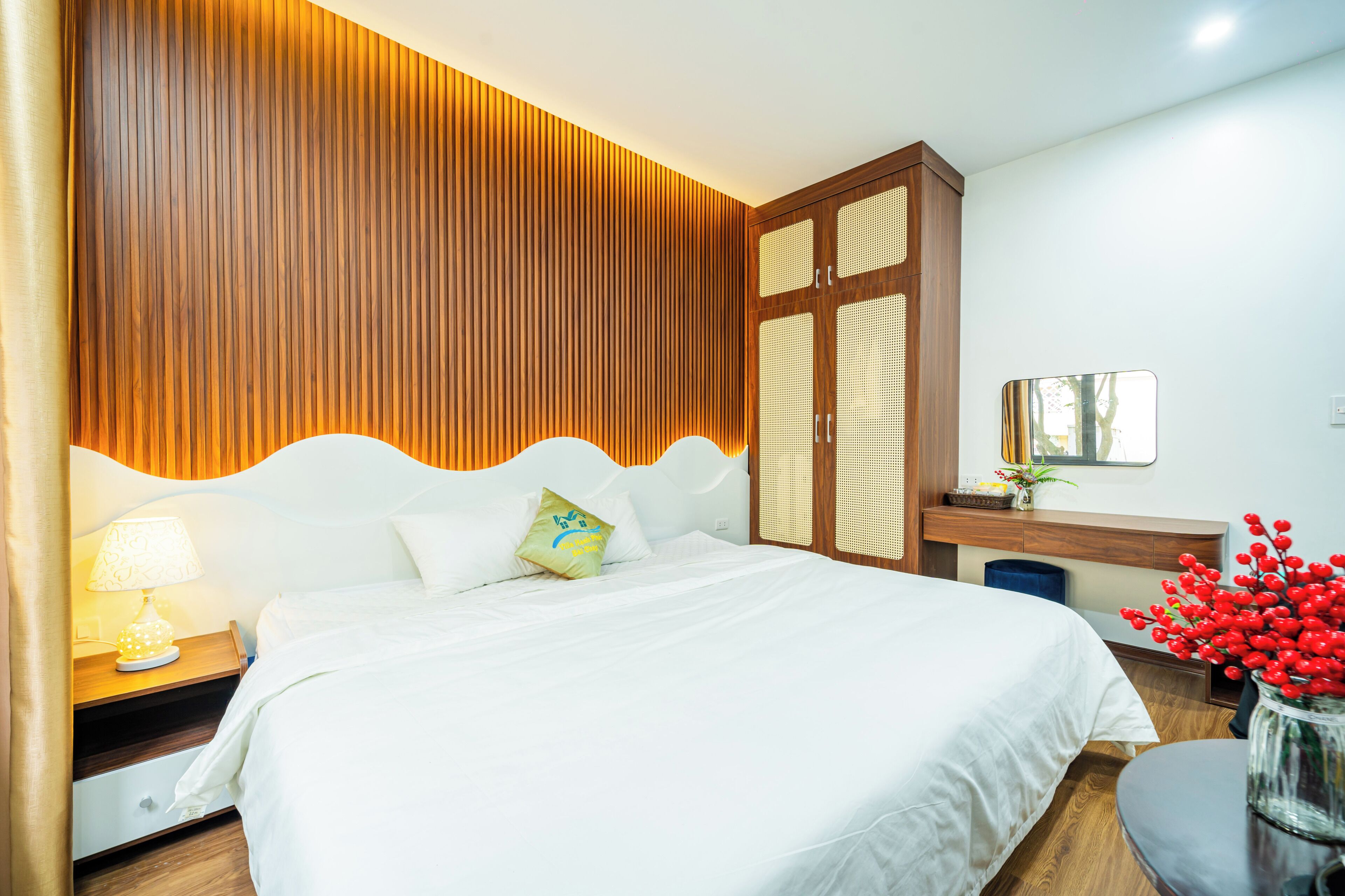 Een hotelkamer met een groot bed, een houten hoofdeinde, een nachtkastje met een lamp, een kleine tafel met een vaas rode bloemen en een spiegel aan de muur.