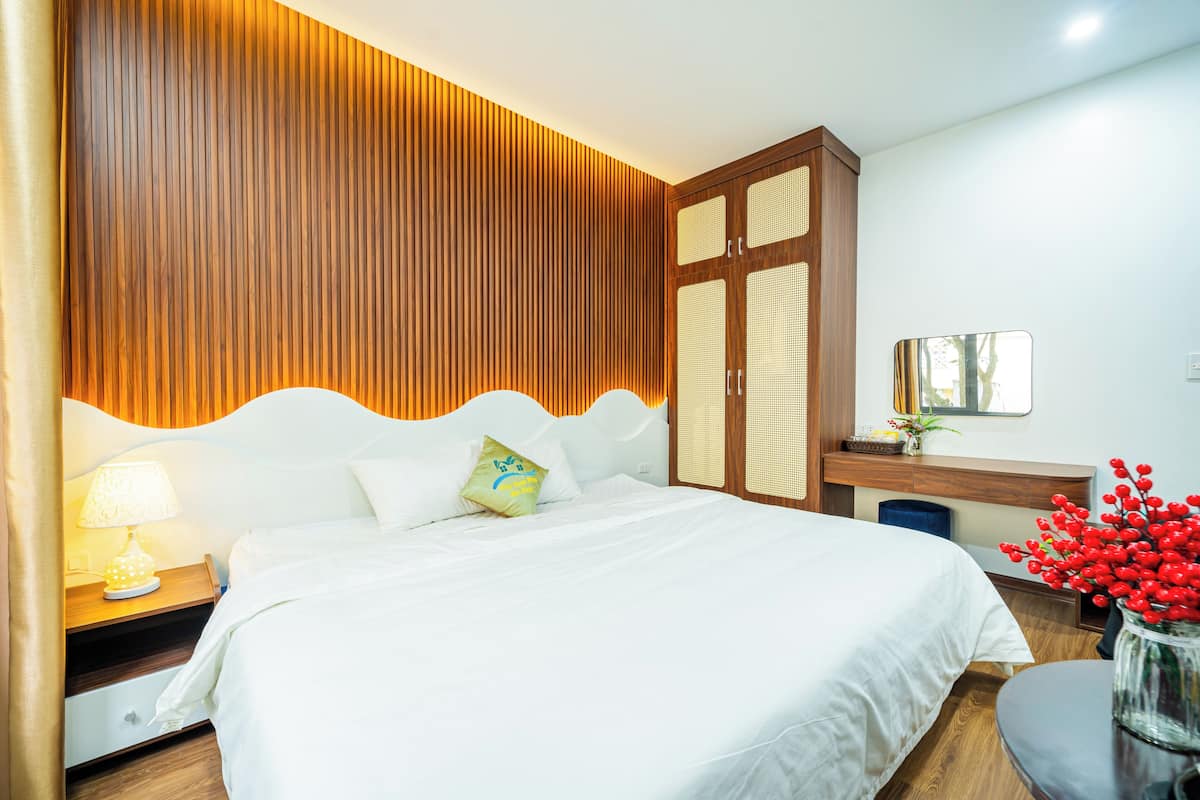 Une chambre d’hôtel avec un grand lit, une tête de lit en bois, une table de chevet avec une lampe, une petite table sur laquelle est posé un vase de fleurs rouges, et un miroir au mur.