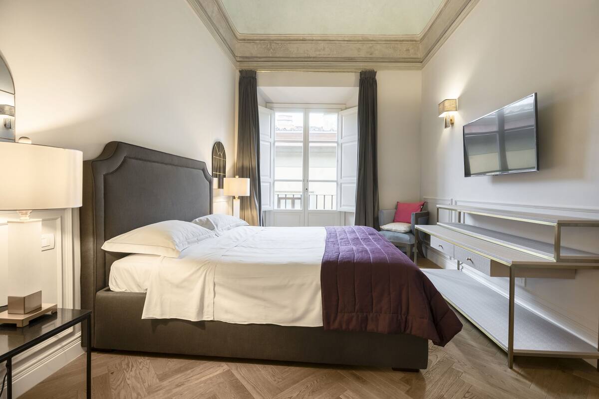 A hotel room with a bed, bedside table, lamp, TV, and a view of the outdoors.