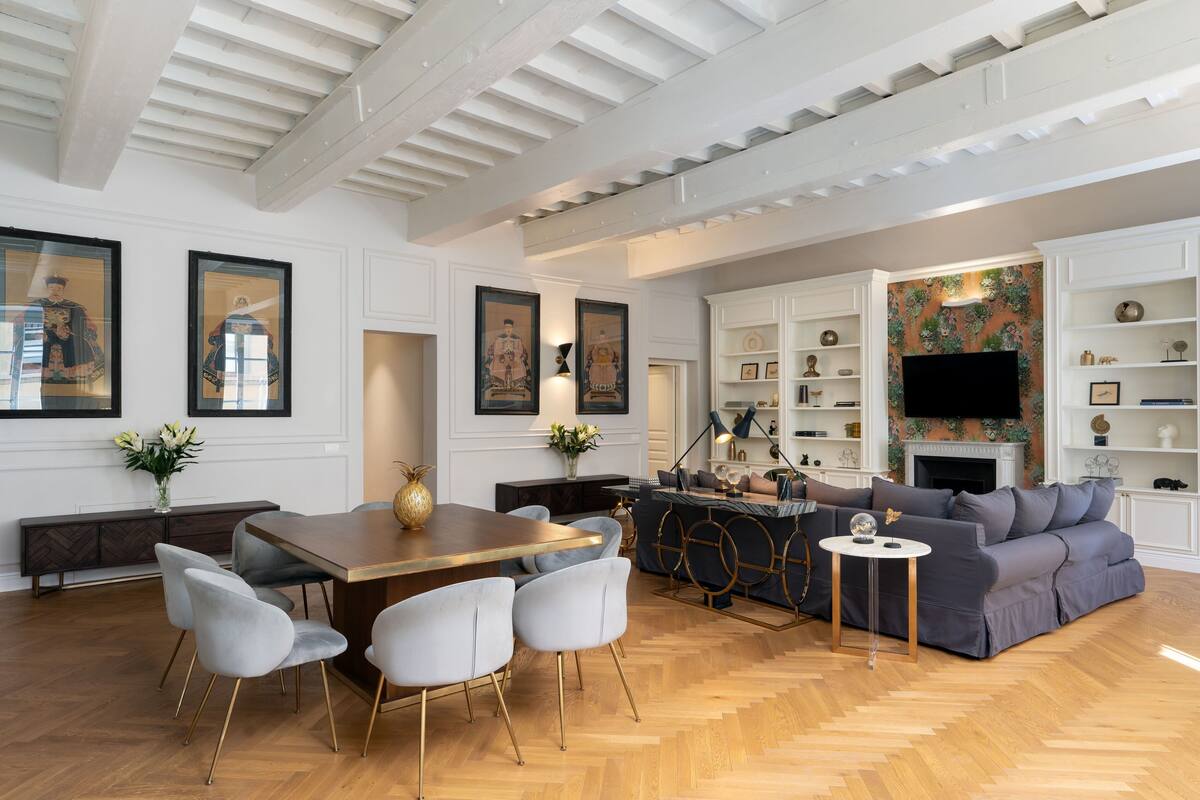 A spacious living room with a wooden herringbone floor, a dining area with a wooden table and modern chairs, a grey sofa, and a fireplace with decorative items.