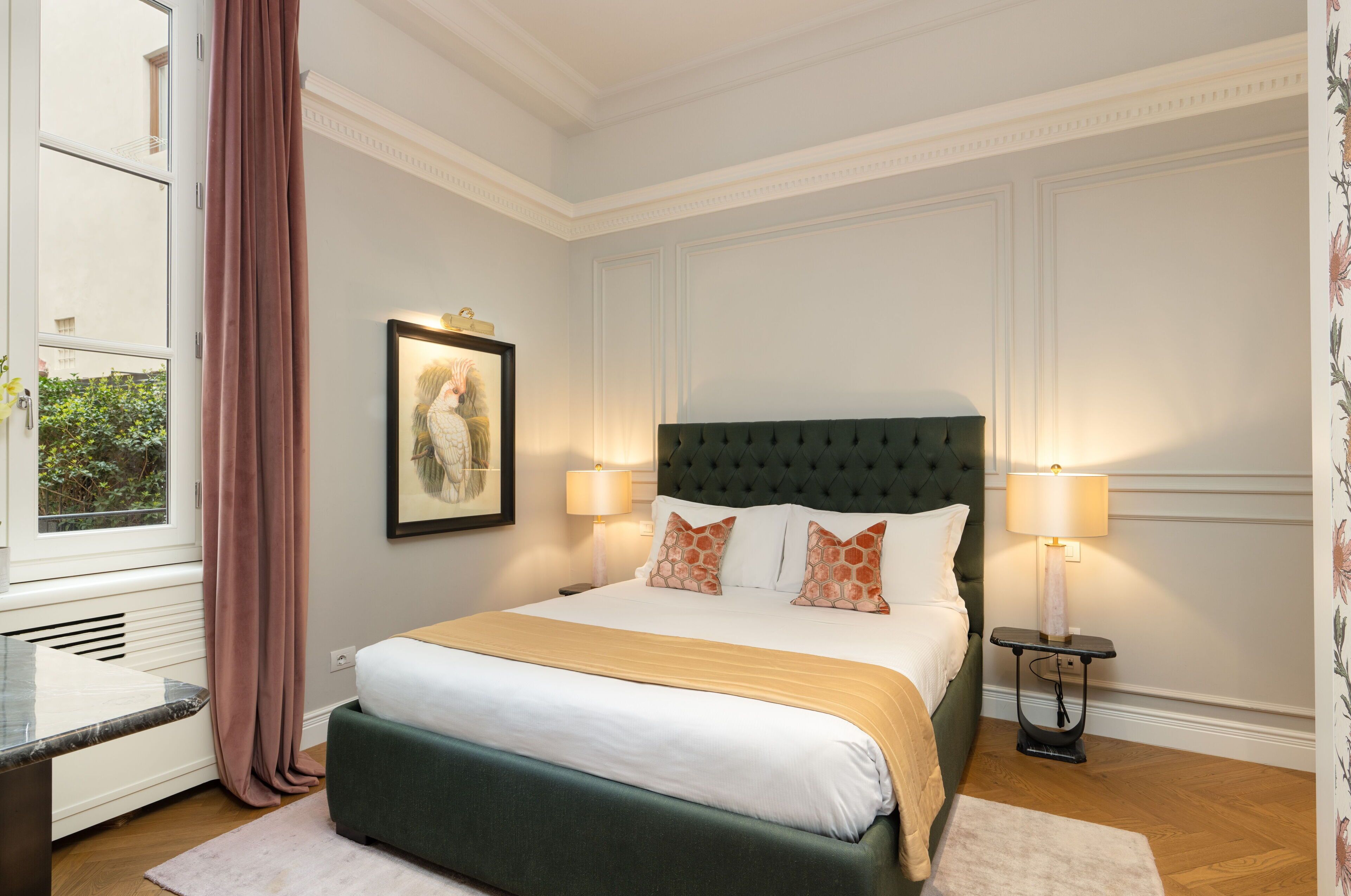Une chambre à coucher bien rangée, avec un grand lit, deux lampes de chevet, un tableau encadré au mur et une fenêtre avec des rideaux.
