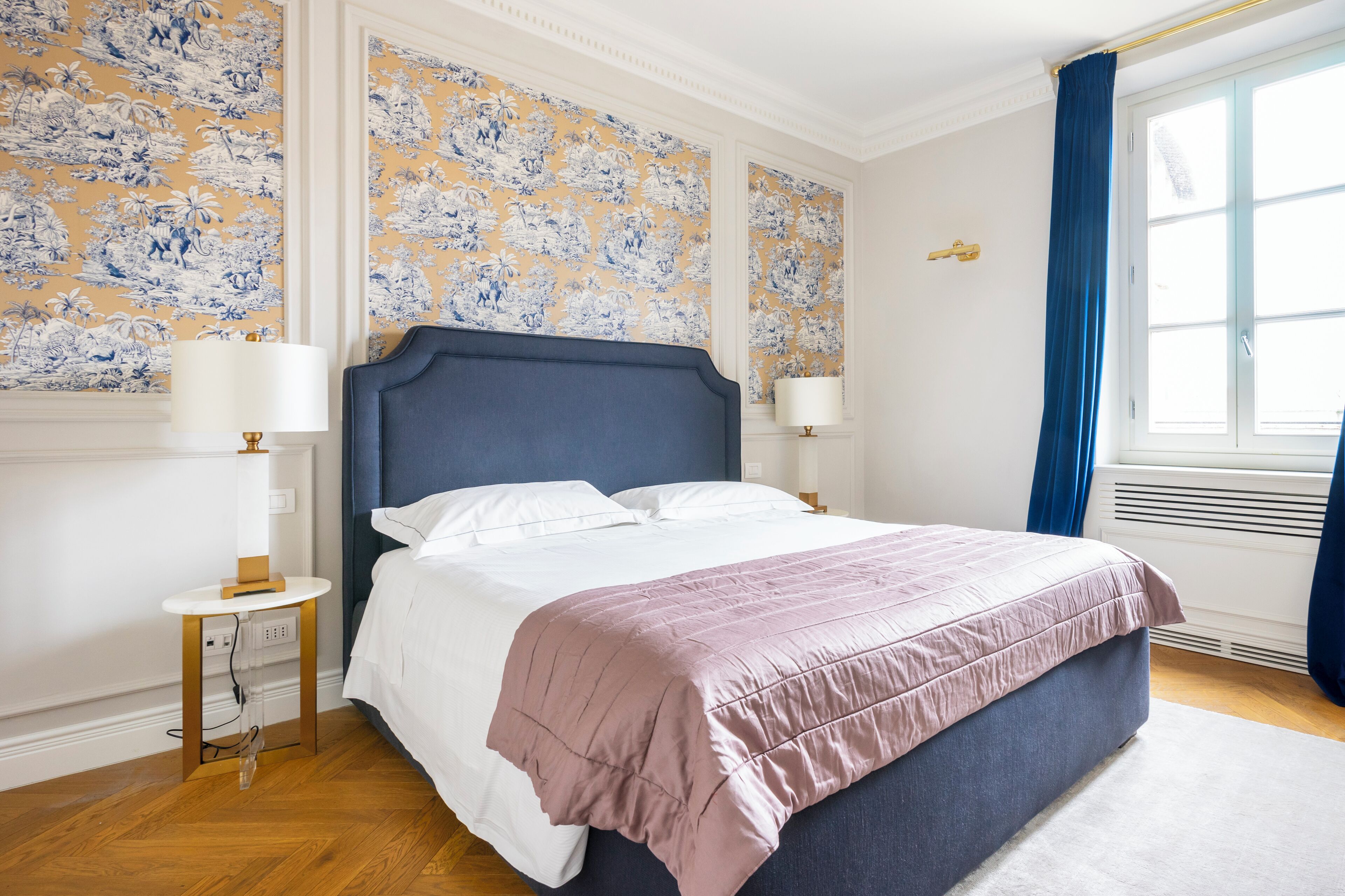 Un dormitorio con una cama grande, dos mesitas de noche, una lámpara auxiliar y una ventana con cortinas azules, además de papel tapiz con motivos florales.
