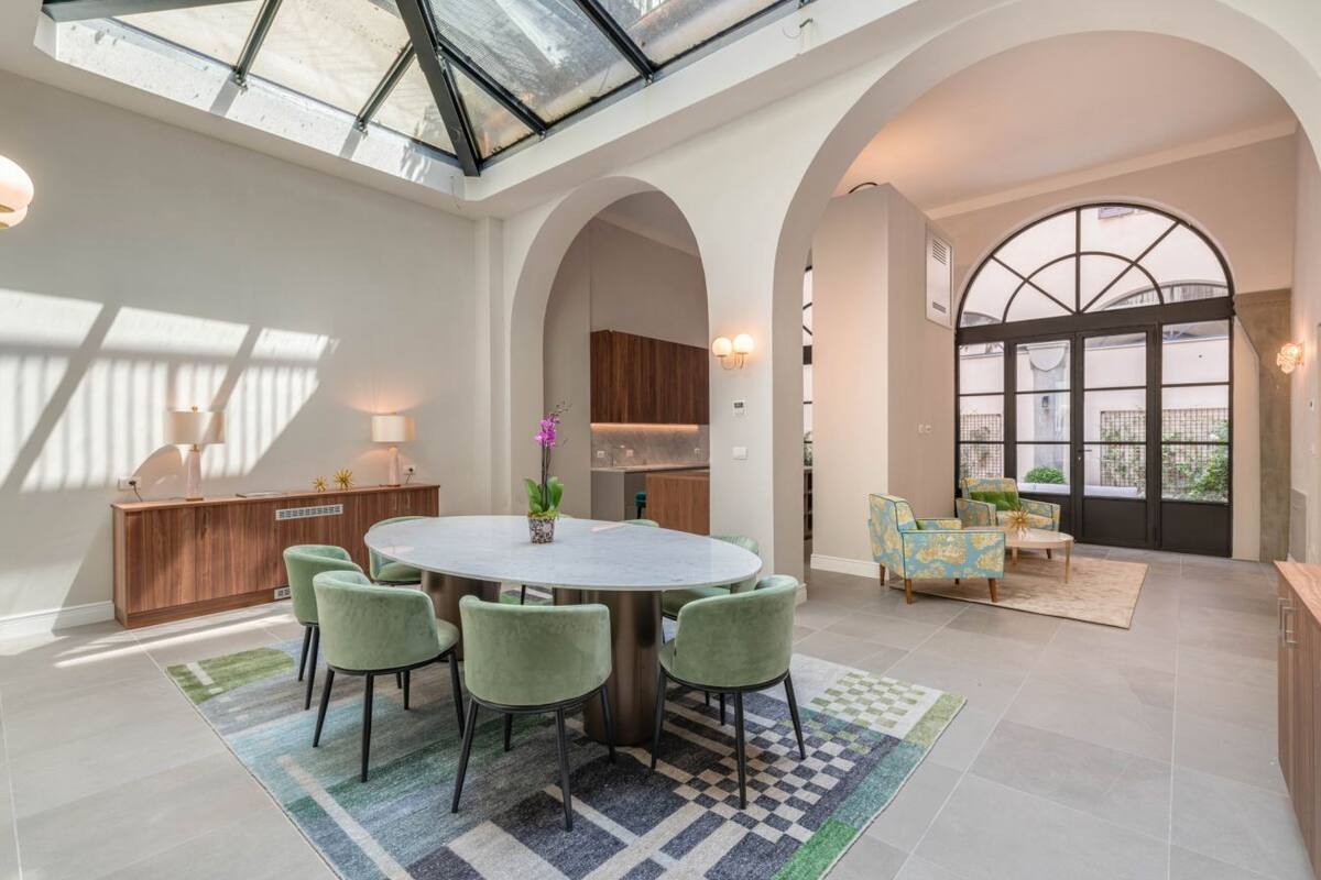 A spacious dining area with a round table and green chairs, a kitchen in the background, and a large arched window with a view.