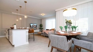 Eine moderne Küche mit einer zentralen Kochinsel, einem Essbereich mit rundem Tisch und grauen Polsterstühlen sowie einem Wohnbereich mit Sofa und Fernseher.