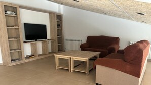 A modern living room with a flat-screen TV, wooden shelving, a brown sofa, and a small coffee table. - Albergue El Muro (Monforte de Moyuela)
