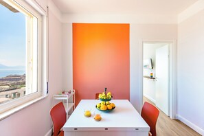 A modern room with a white table, orange wall, and a view of the city. - Napoli Bellavista Apartment (Naples)