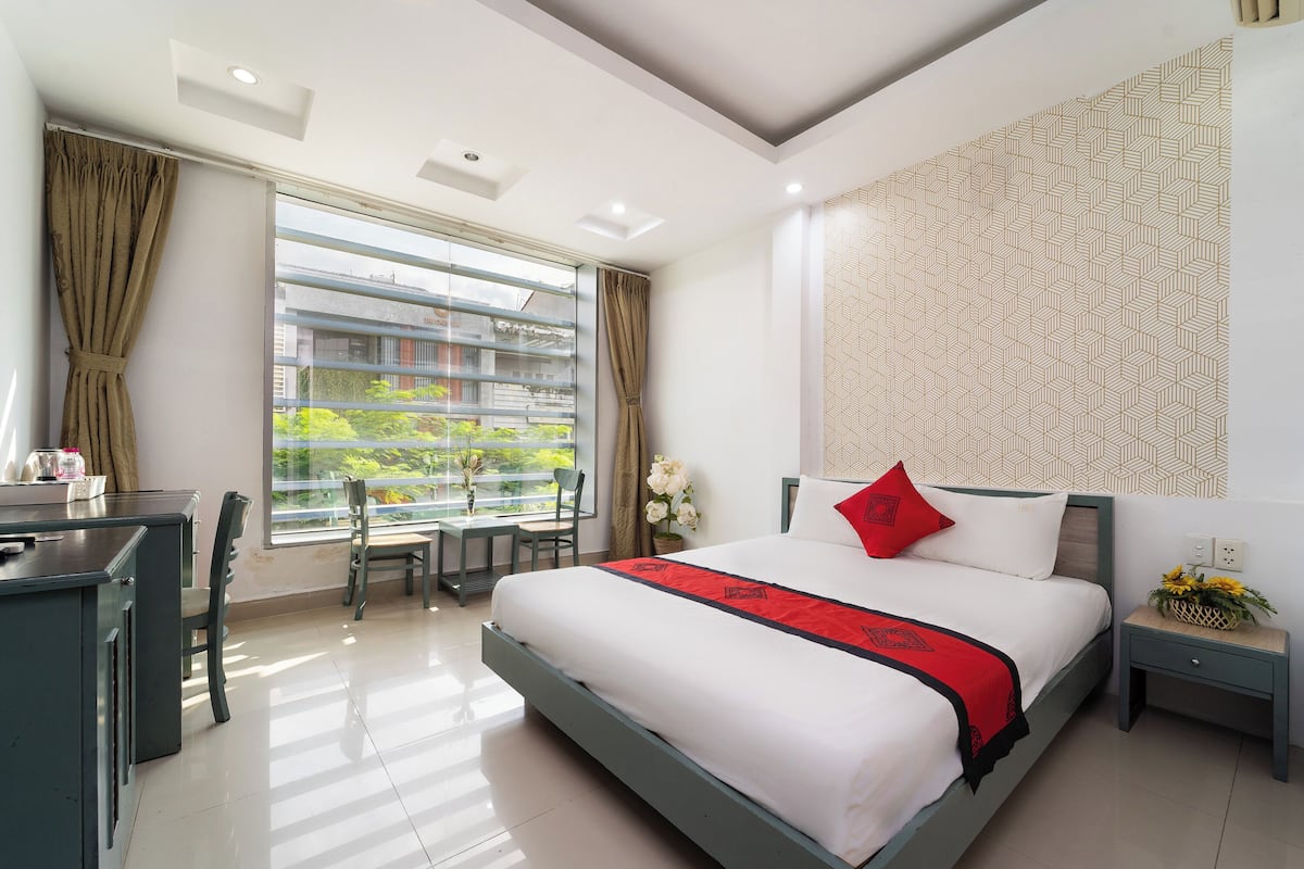 A hotel room with a bed, desk, chairs, and a view of greenery outside.