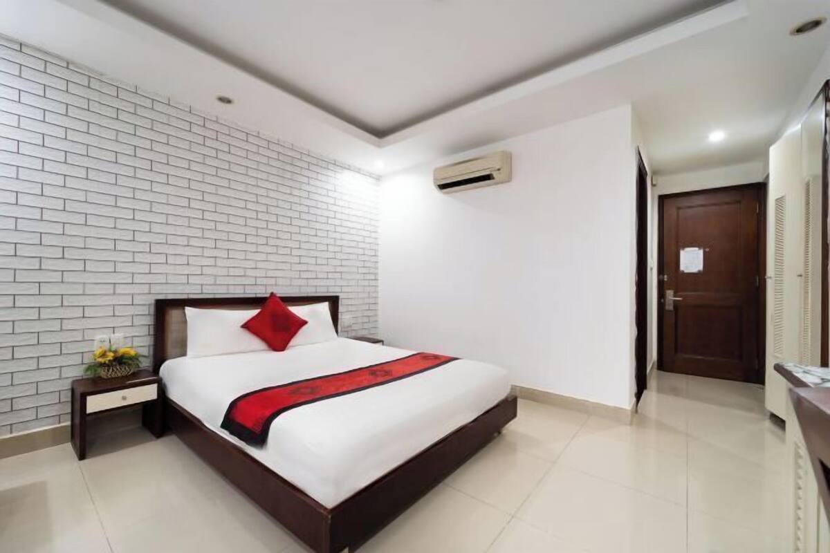A hotel room with a brick-patterned wall, a wooden bed frame, a red pillow, a white bedspread, and a small bedside table with a vase of flowers.