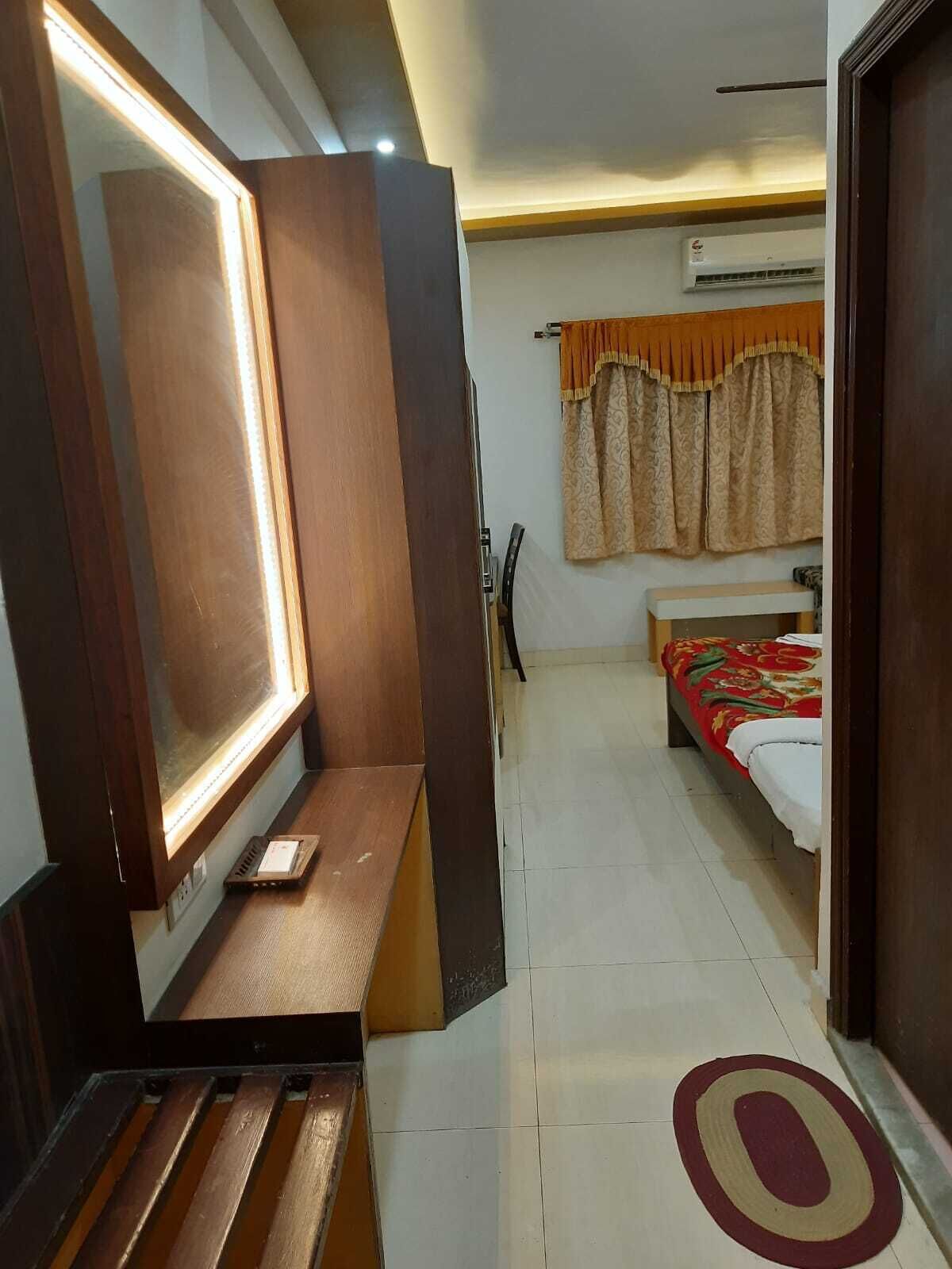 A hotel room with a wooden bench, a large mirror, a bed with a red and white bedspread, and a window with patterned curtains.