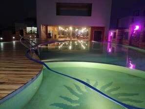 A swimming pool with a wooden deck and a glass wall reflecting lights. - Hotel Sunshine (Baharampur)